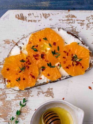 Orange slices with thyme and chilli flakes on a slice of ricotta toast that has a honey vinaigrette drizzled over.