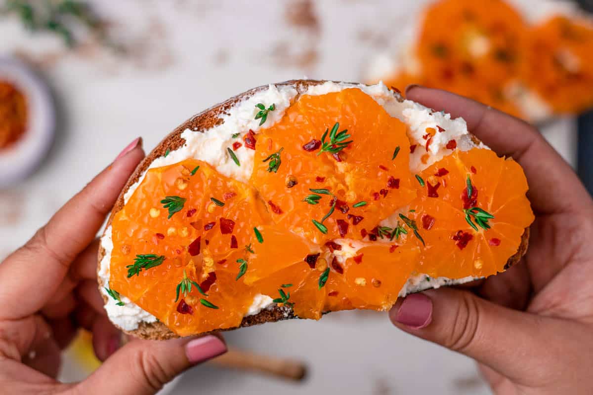 A slice of orange & honey ricotta toast being held by two hands.