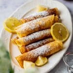 Pancakes made using Queen Elizabeth's royal pancake recipe, rolled, and on a white dish with lemon wedges and sugar.