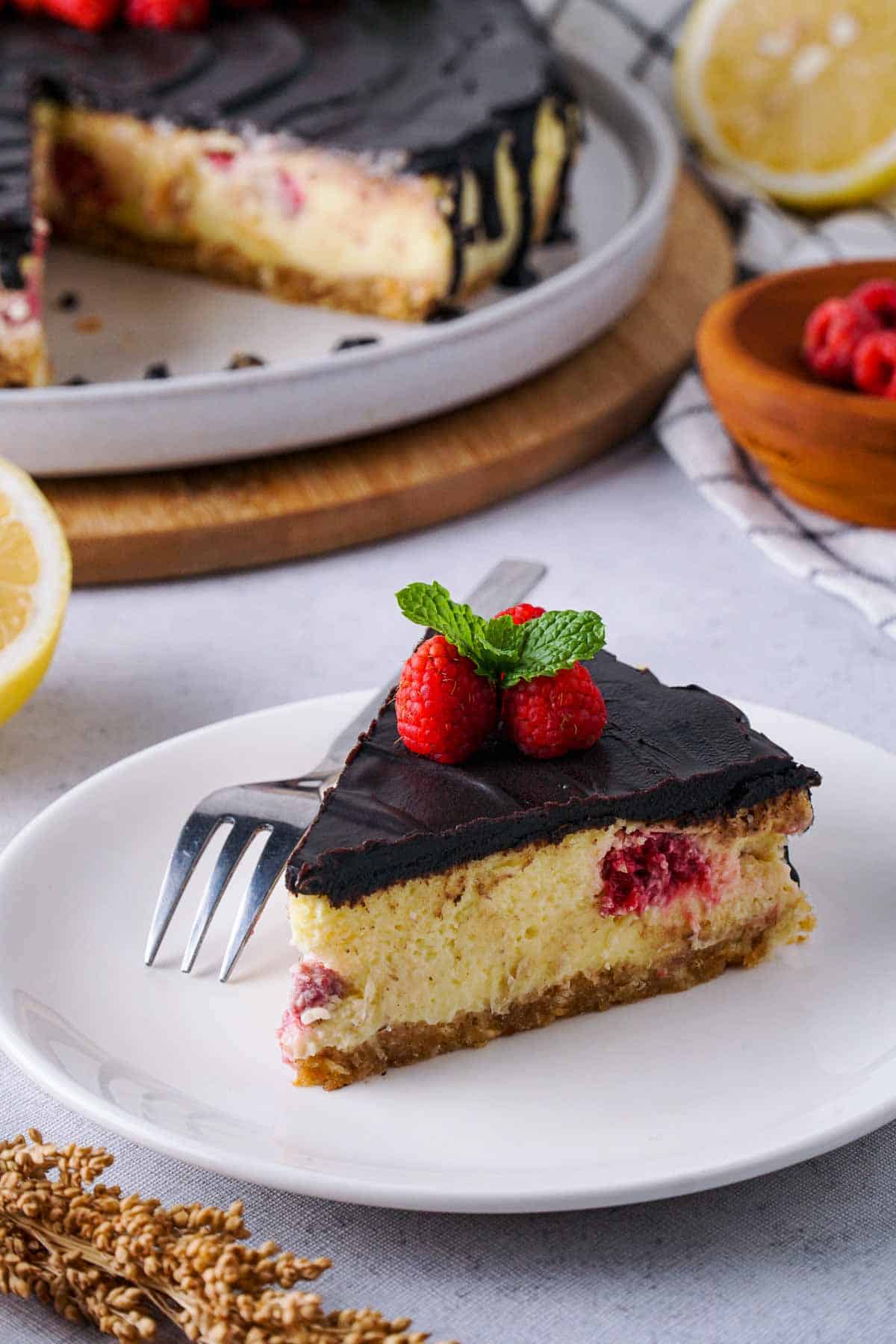 A slice of cheesecake on a plate. It is covered with chocolate ganache and filled with raspberries. In the background, some ingredients and more cake can be seen.