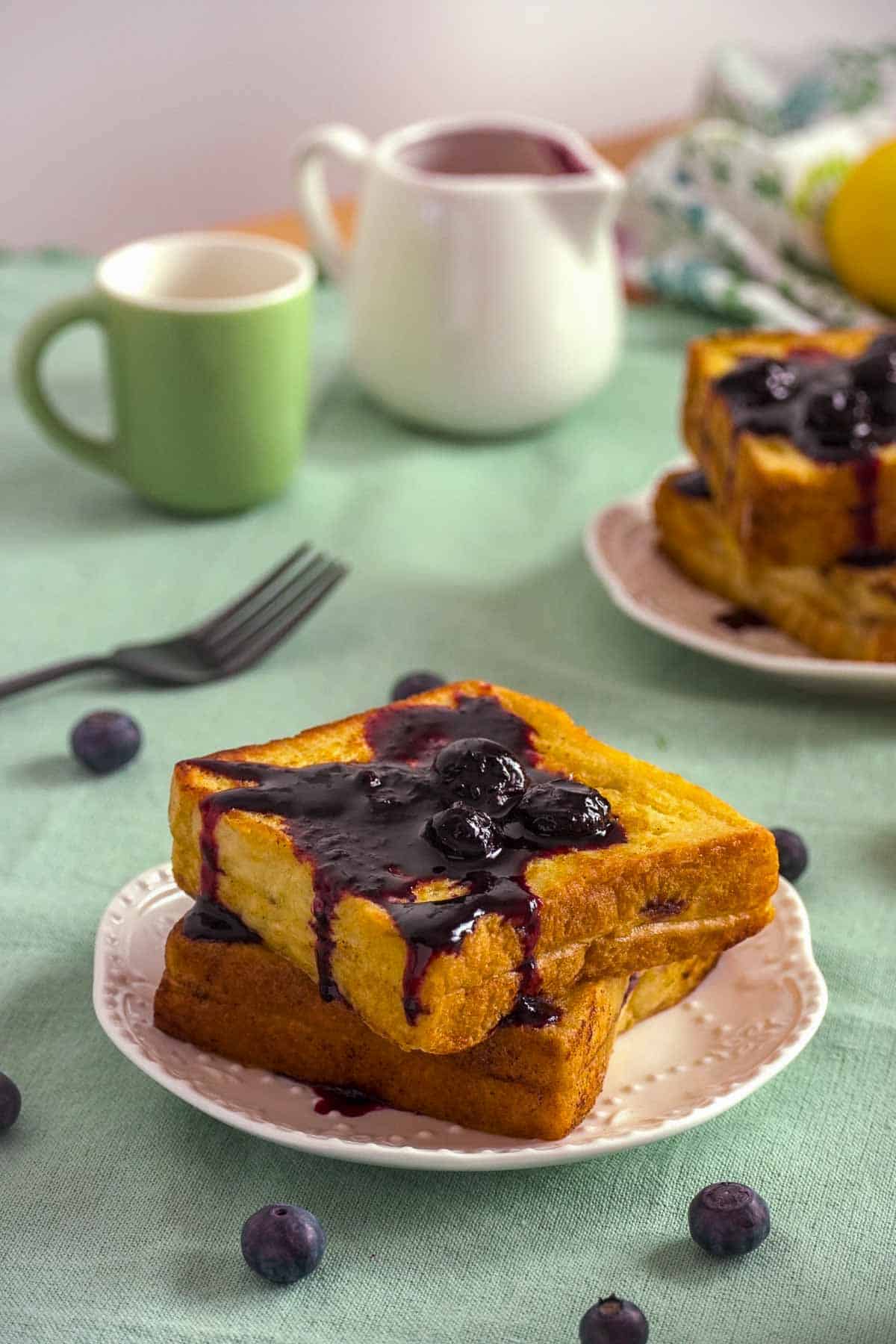 A stack of Lemon Blueberry French Toast on a plate with a green tablecloth and some props in the background.