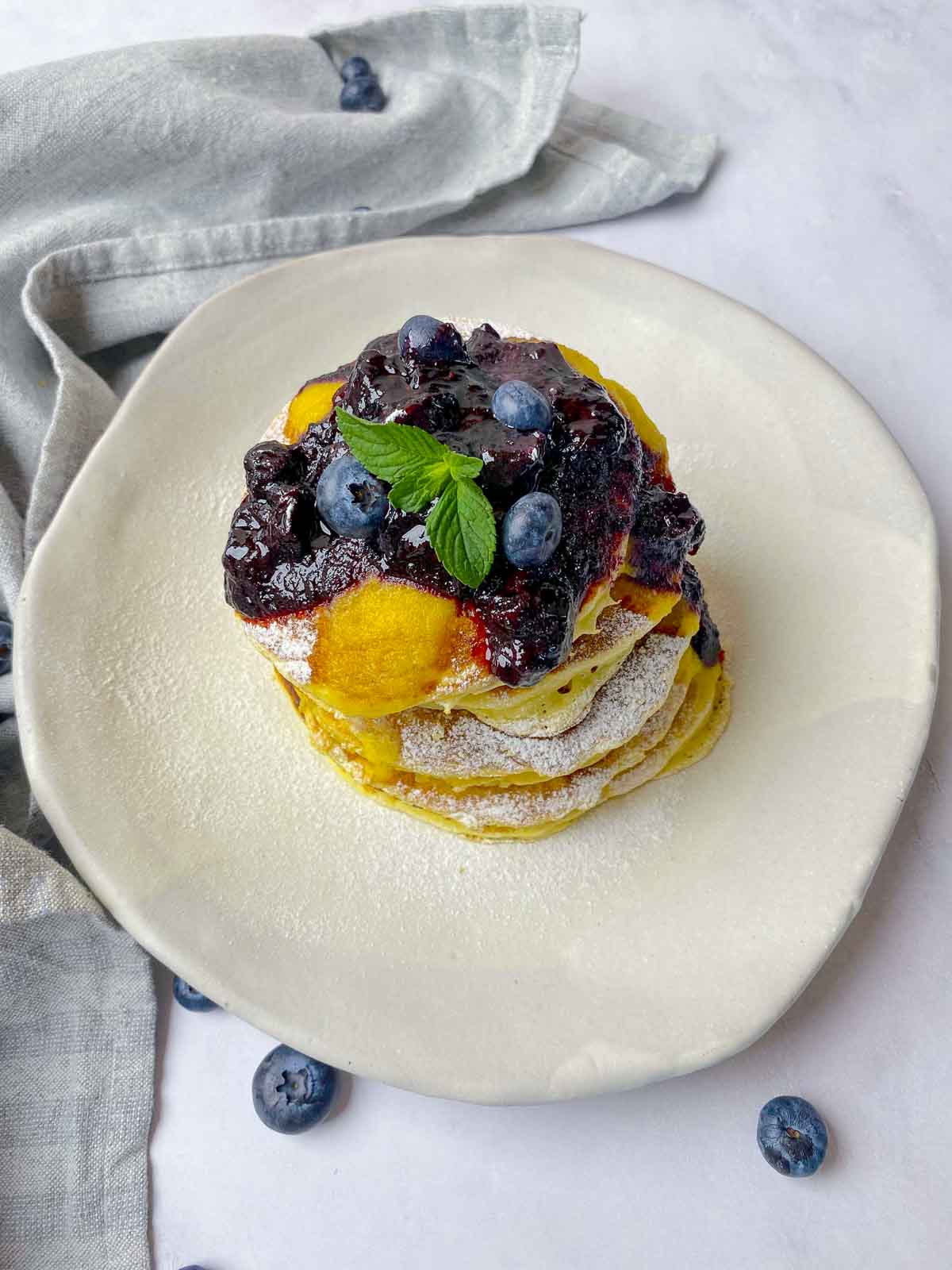 A plate of lemon ricotta pancakes covered in blueberry compote with some fresh blueberries added for good measure.