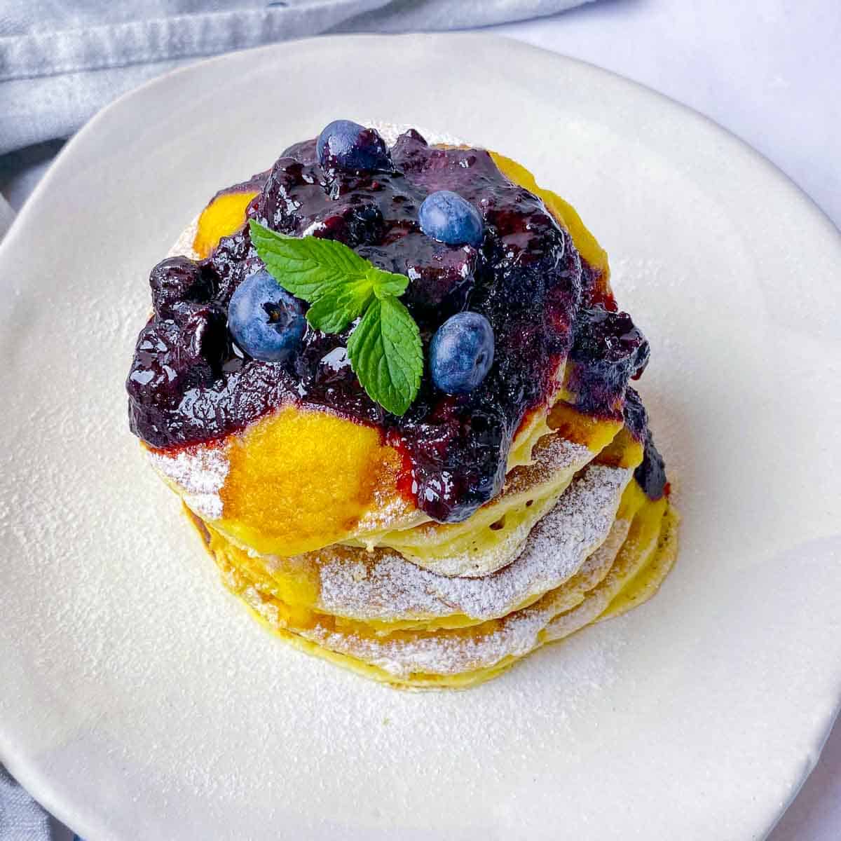 A stack of fluffy lemon ricotta pancakes on a white plate, garnished with blueberry compote.