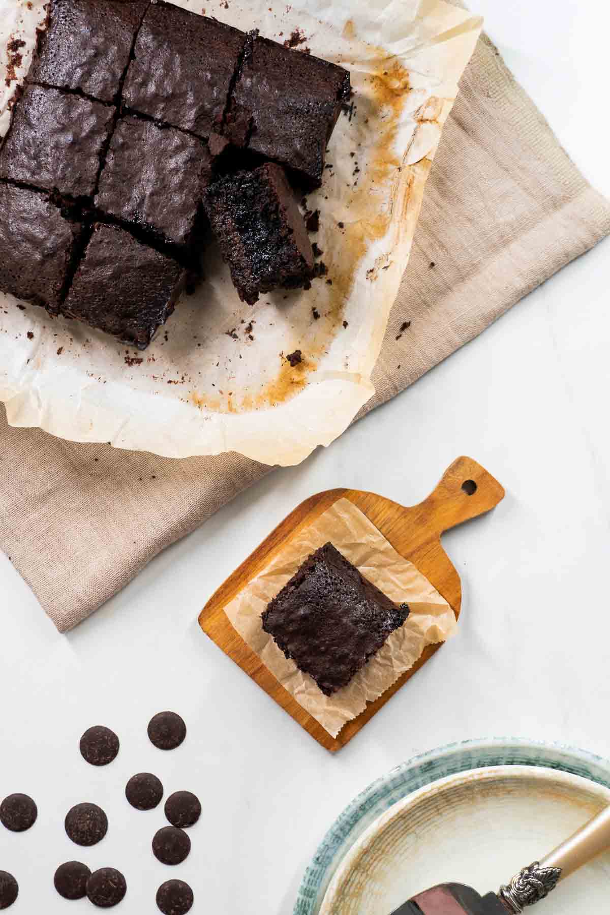 A flatlay showing the full batch of baked brownies, as well as a single piece that's on a sheet of parchment paper.