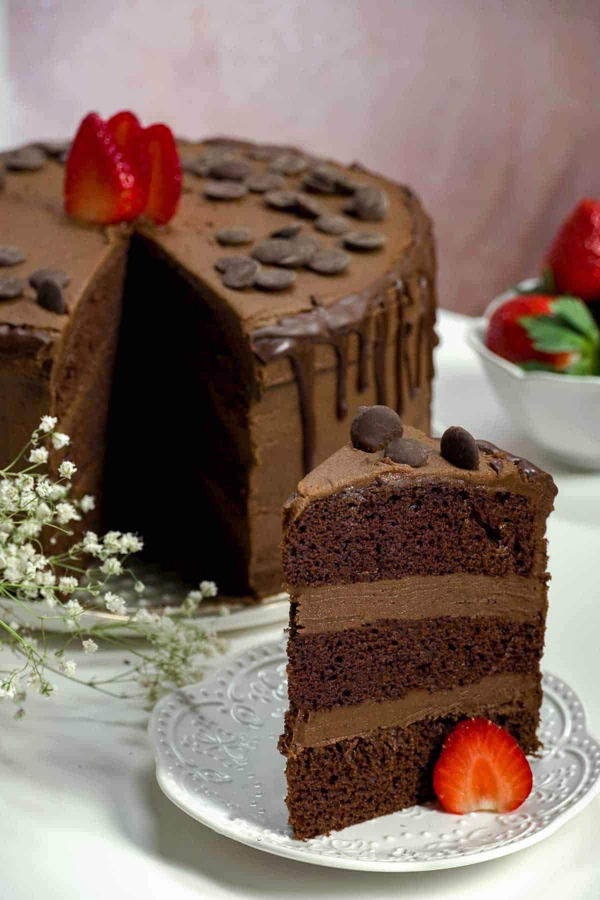 A slice of chocolate cake on plate with a strawberry. The remainder of the cake can be seen in the background.