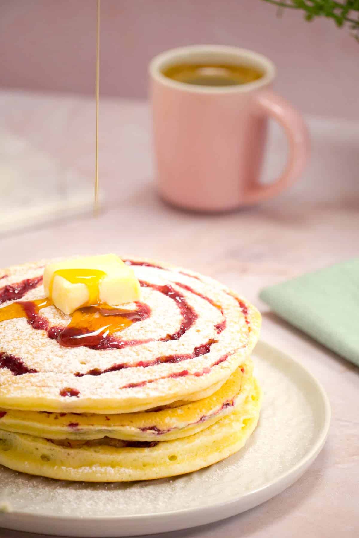 Raspberry swirl pancakes with icing sugar having maple syrup poured on to them.