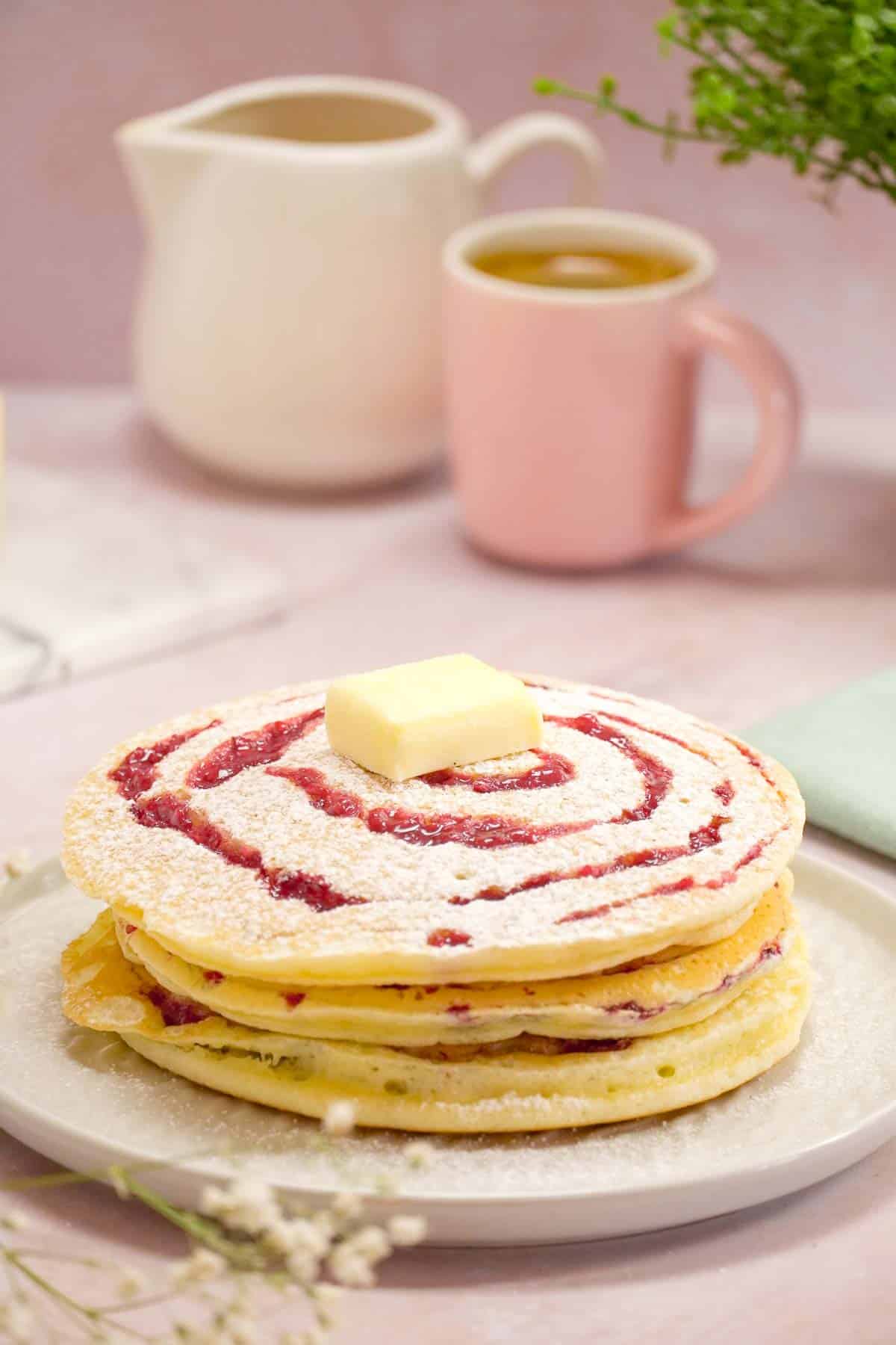 Three raspberry swirl pancakes on a white plate. There are a decorative mug and jar in the background.