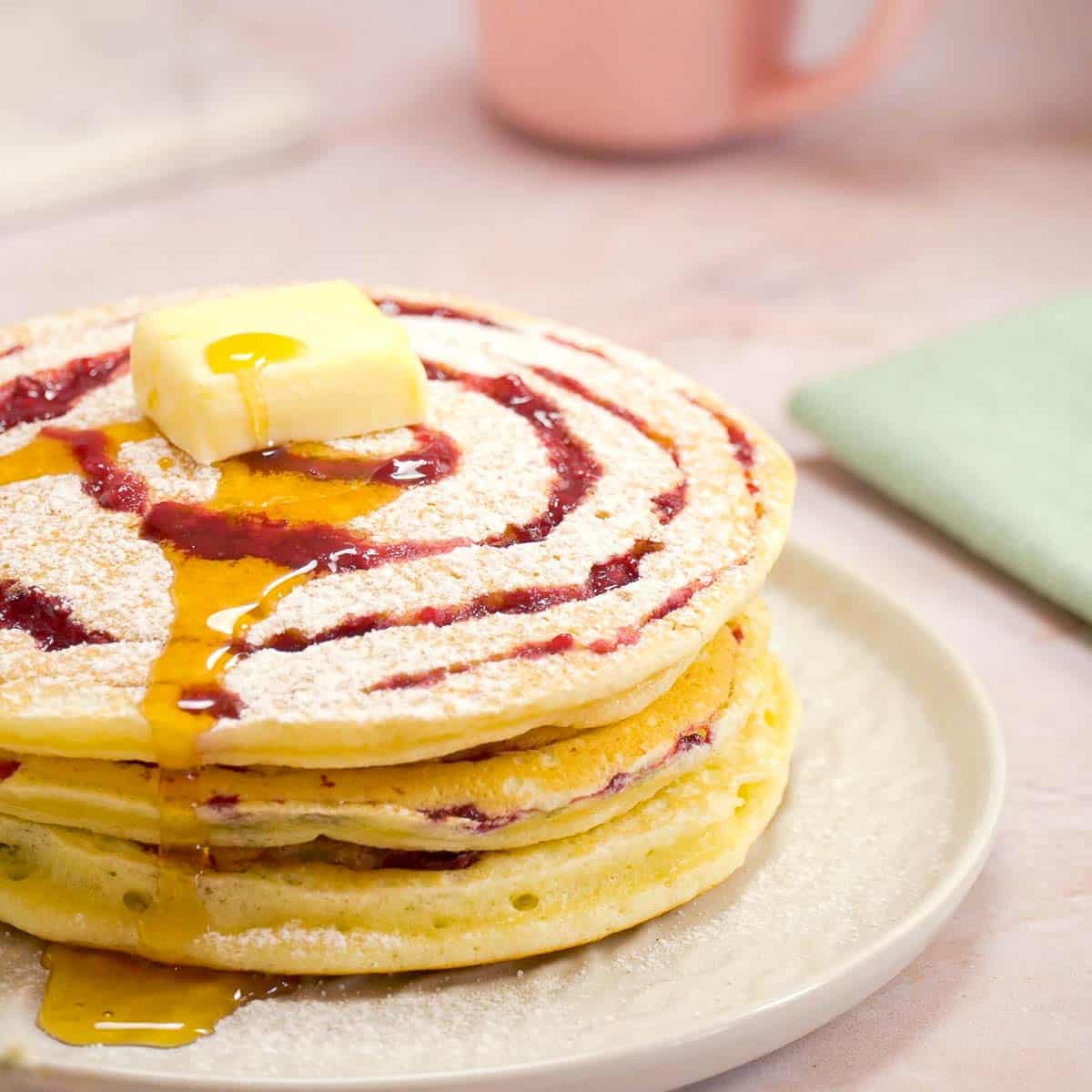 Maple syrup dripping off the side of a stack of raspberry pancakes with a swirl pattern on top.