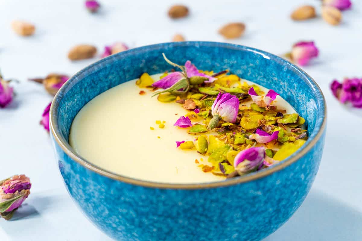 A close-up shot of a blue bowl of mahalabia. The rose petals and pistachios provide a pop of colour against the white pudding.