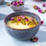 A purple bowl filled with chilled mahalabia, garnished with crushed pistachios and rose petals.