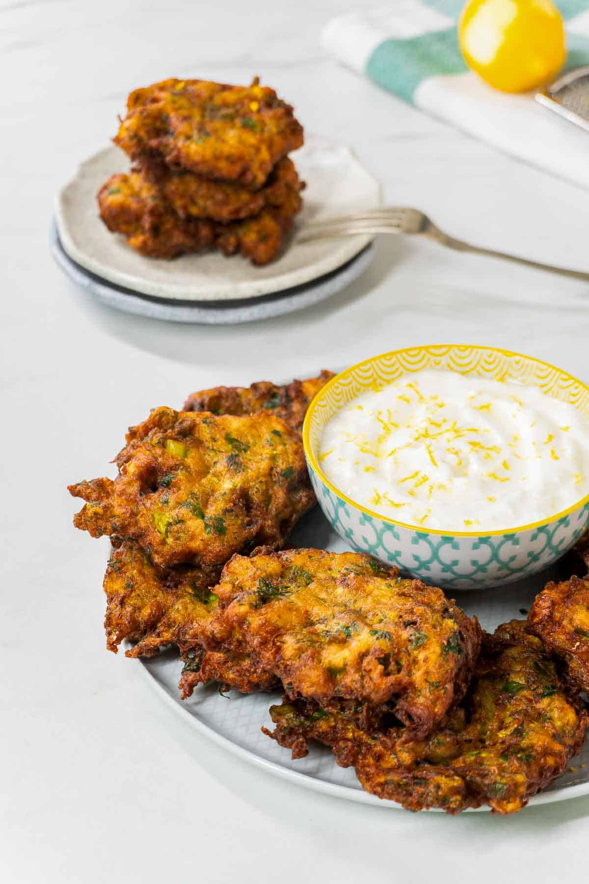 Mucver surrounding a bowl of yoghurt dip. More individual zucchini fritters are visible in the background.