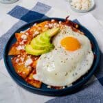 A blue plate filled with chilaquiles rojos, garnished with a fried egg, queso fresco, and avocado slices.