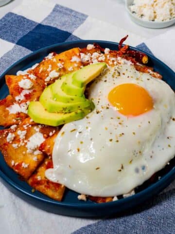 A blue plate filled with chilaquiles rojos, garnished with a fried egg, queso fresco, and avocado slices.