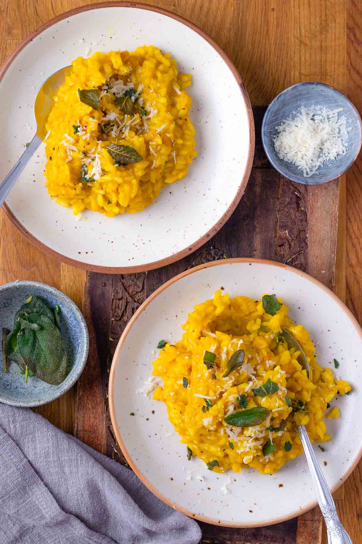 A bird's eye shot of two plates of creamy pumpkin risotto, garnished with sage. There is some shredded cheese in a bowl next to the top plate.