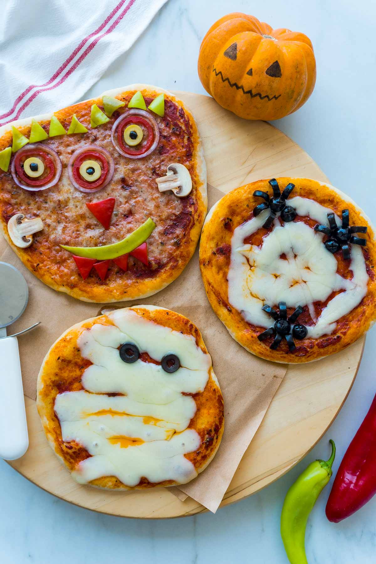 Three halloween pizzas on a wooden board with a pizza slicer and a pumpkin next to them.