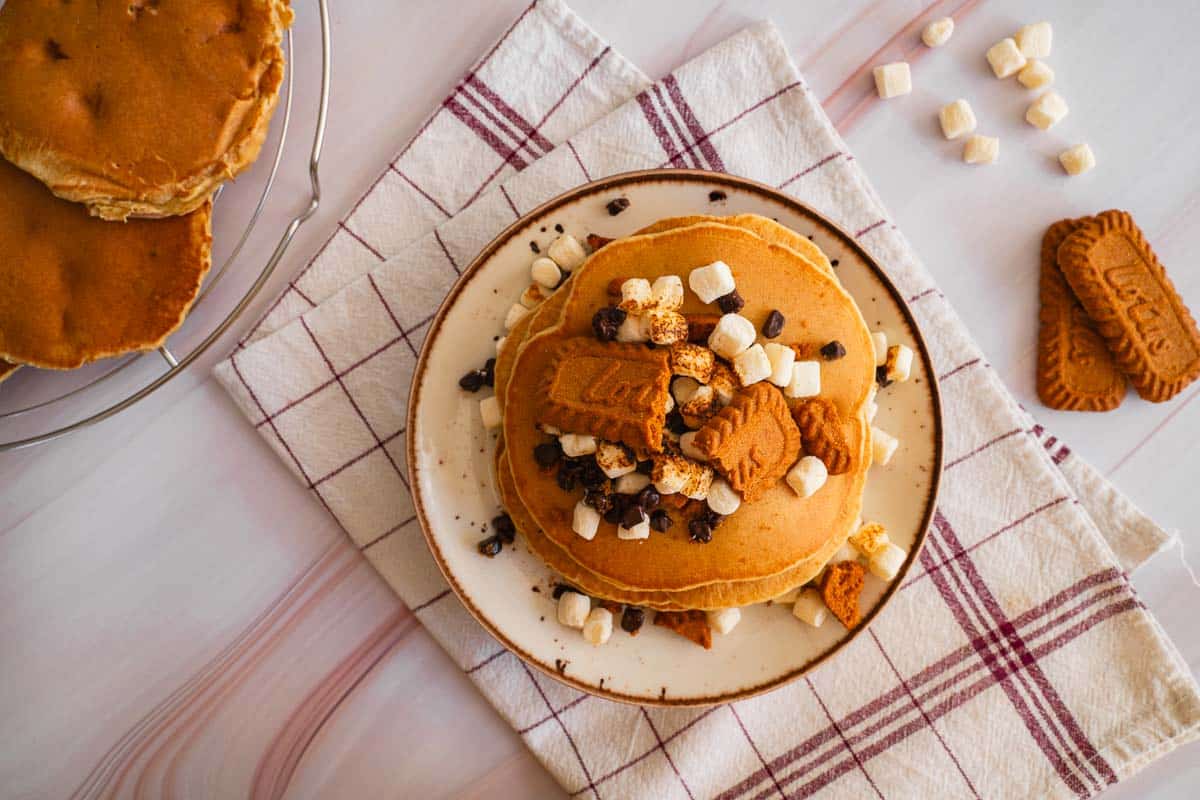 A bird's eye view of a plate of pancakes. The crumbled Lotus Biscoff cookie is clearly visible with marshmallows and chocolate chips below it.