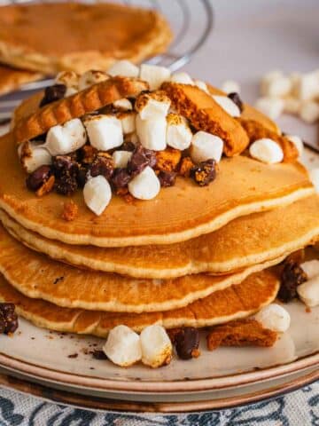 A large stack of four S'mores Pancakes. The pancakes are topped with a generous portion of torched mini marshmallows, crumbled cookies, and chocolate chips.