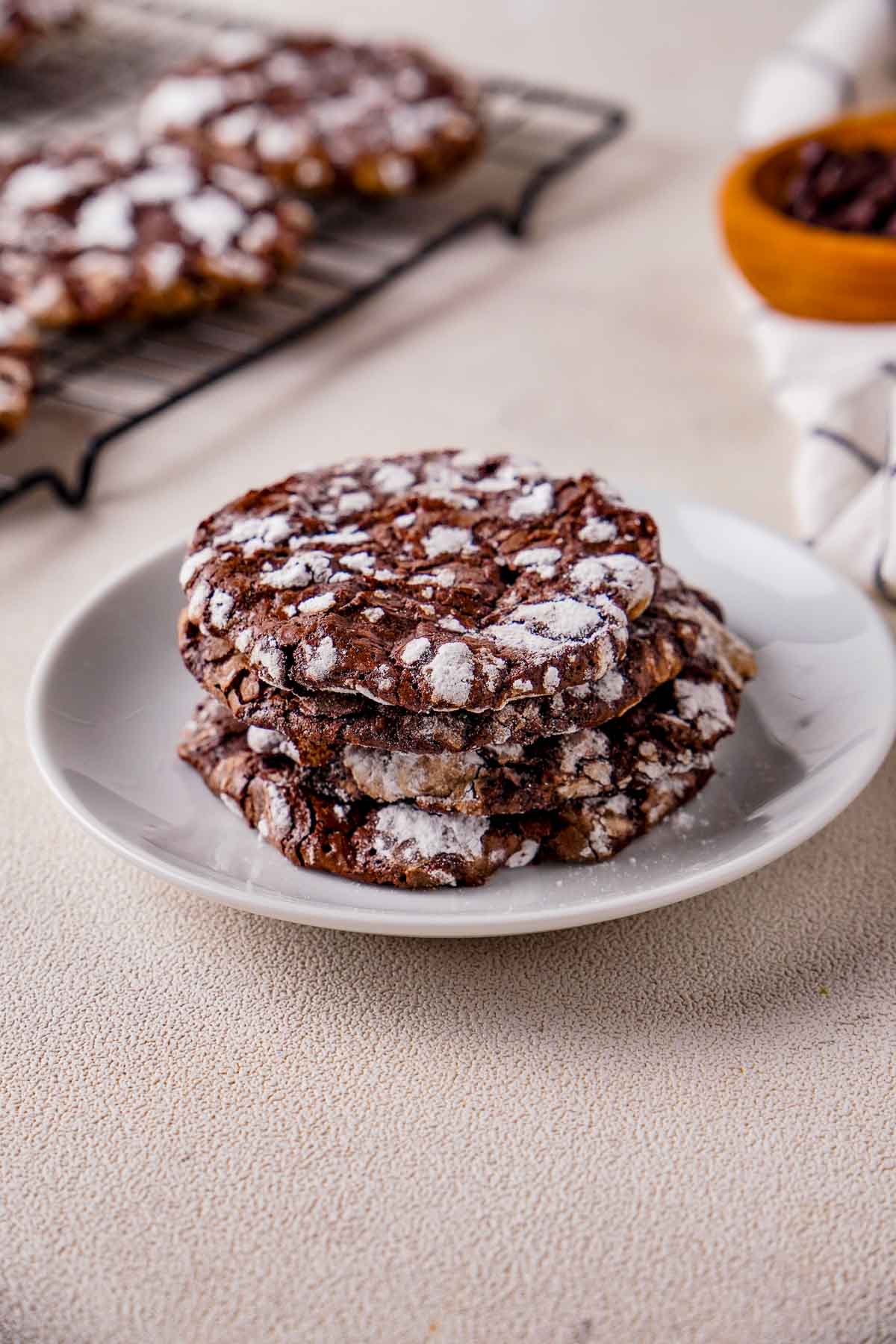 Chocolate crinkle cookies stacked on white plate, dusted with powdered sugar, with cooling rack blurred in background.