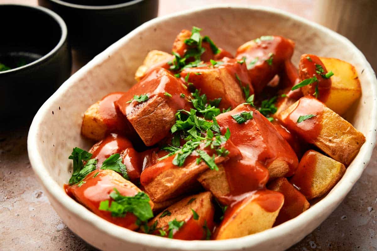 A white bowl of Spanish fried potatoes with bravas sauce and fresh parsley.