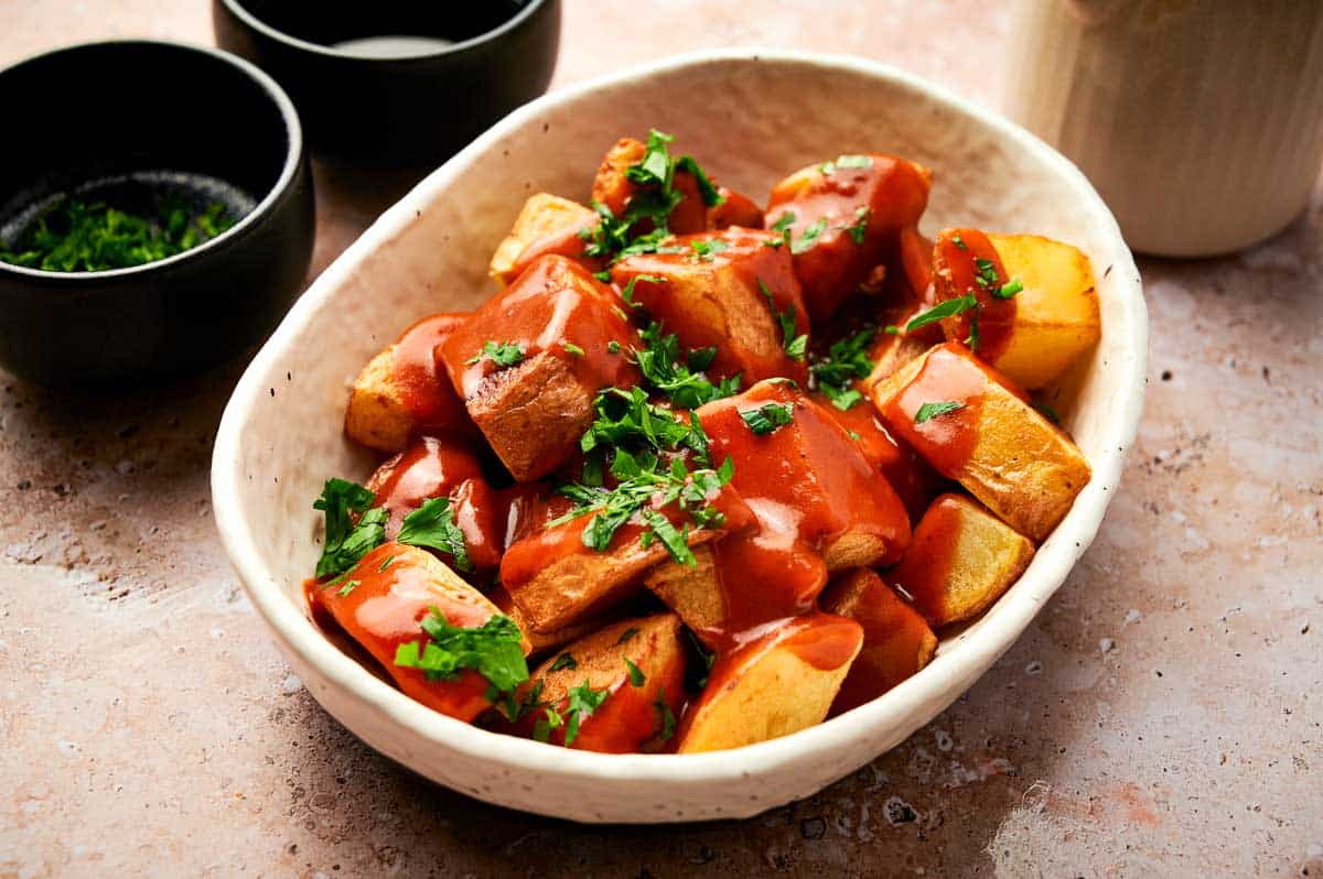 A white bowl of patatas bravas, with two black bowls beside it containing parsley and salt.