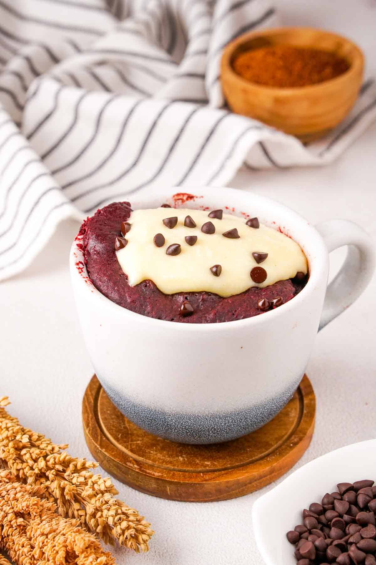 Red velvet cake in a mug on a wooden coaster. Chocolate chips are visible in the bottom corner of the photo.