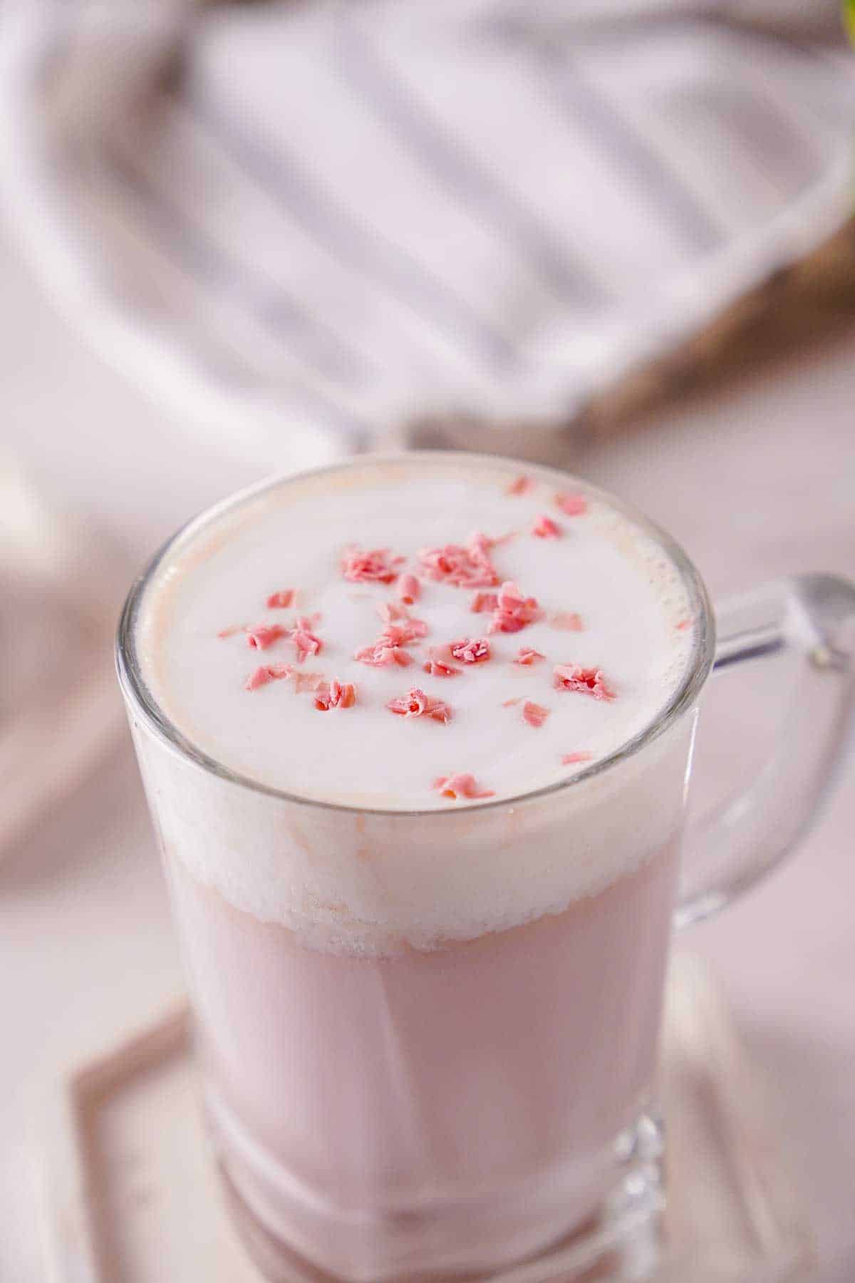 A clear mug of hot chocolate with a pink color. The hot chocolate is garnished with ruby chocolate shavings.