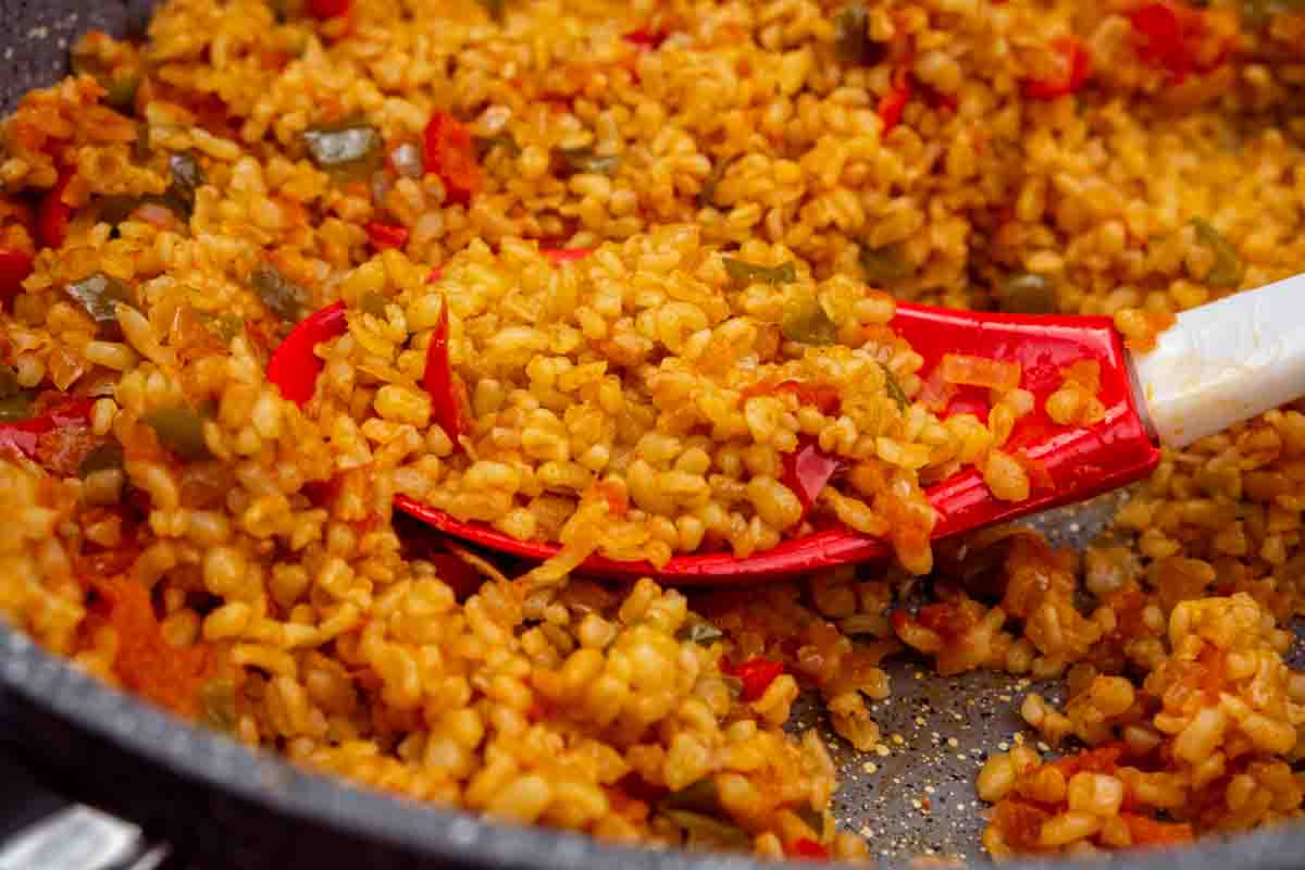 A close-up of bulgur pilaf being cooked in a pan. A red spatula has lifted some of the bulgur.