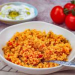 A white bowl of bulgur pilavi with tomatoes. A spoon is digging in to get a bite.