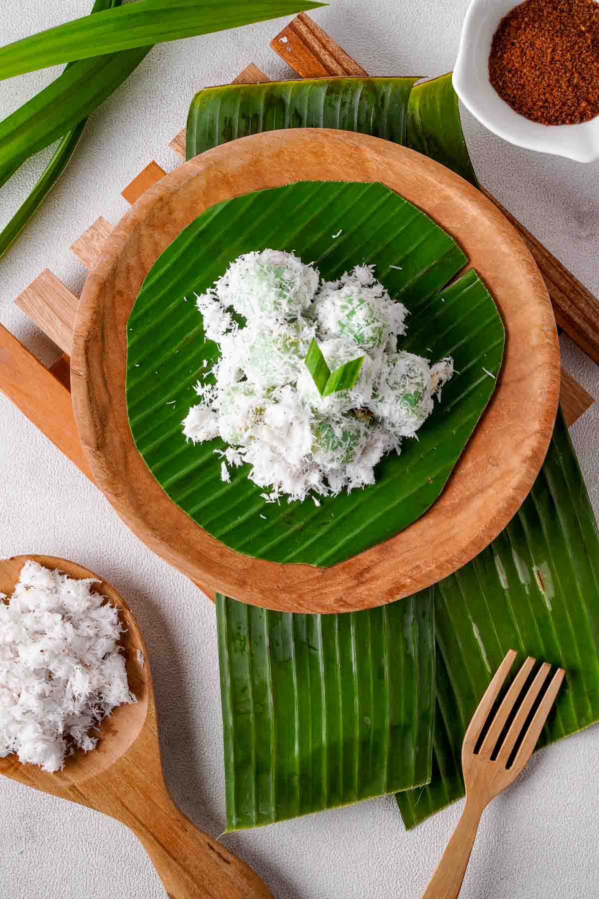 A top down view of several Indonesian klepon, covered in lots of grated coconut, served on a banana leaf. A wooden serving dish with more grated coconut can be seen at the bottom of the image.