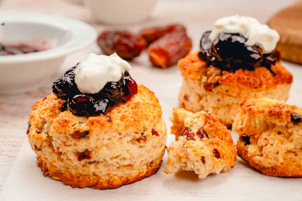 A close-up of two date scones with whipped cream and jam as toppings. Some more dates can be seen in the background.