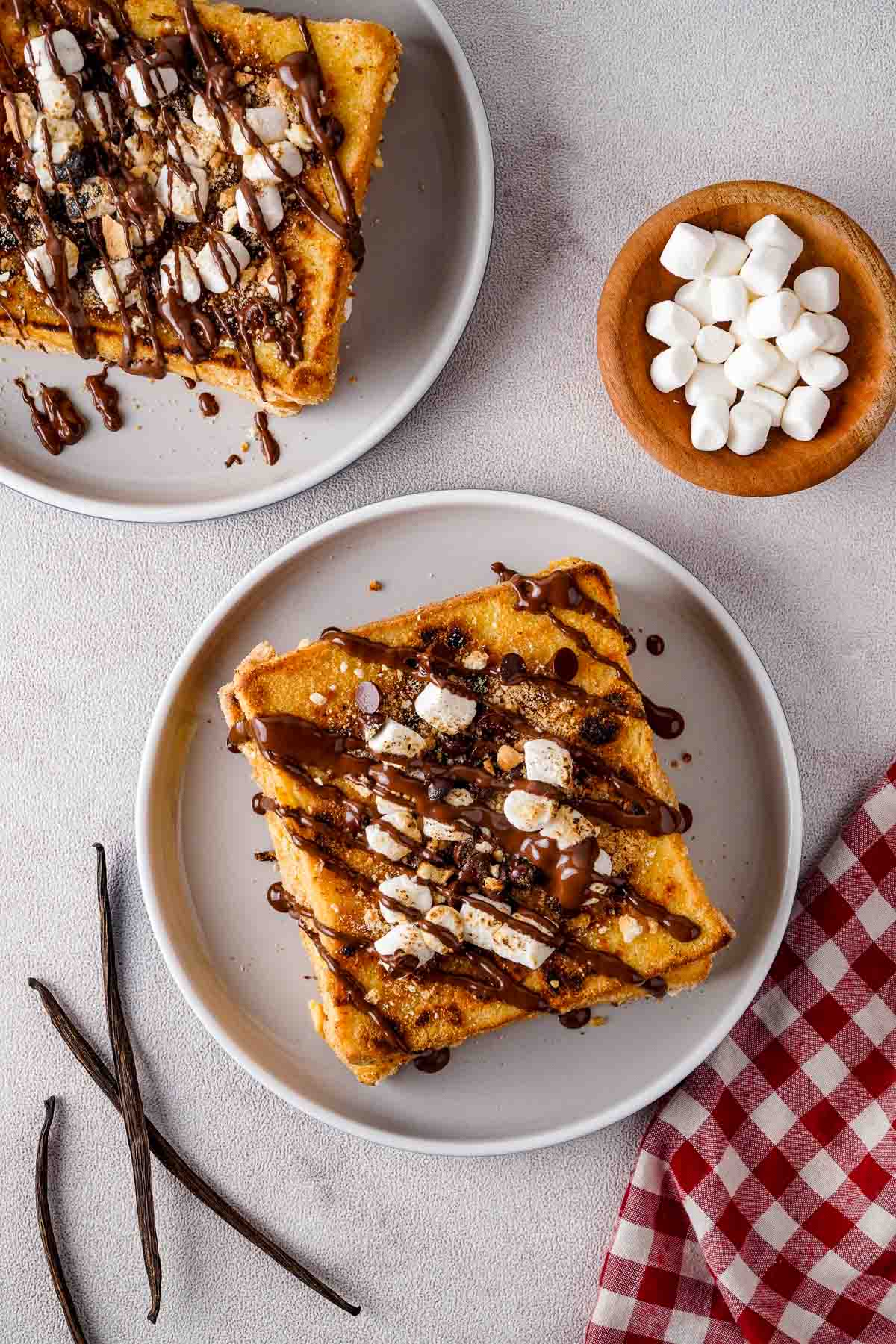 A top down view of two plates of French toast topped in chocolate drizzle, marshmallows, and crushed graham crackers.