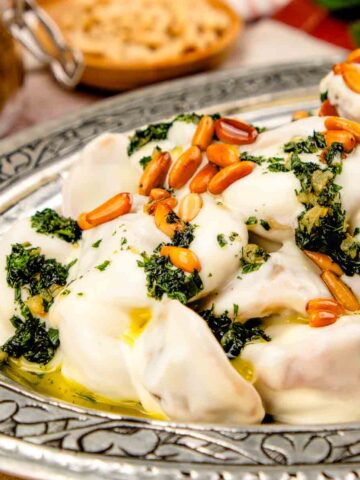 A plate of Middle Eastern Shish Barak dumplings, covered in a garlic butter sauce, and toasted pine nuts.