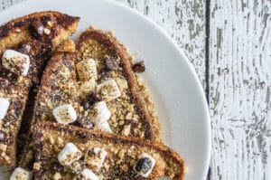 A photo of S'mores French Toast on a white plate atop a rustic white wooden table.