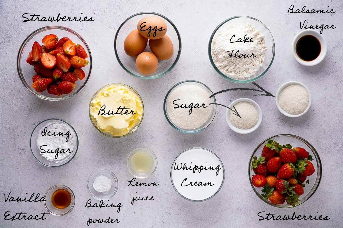 A top-down photo of all the ingredients in bowls. The photo shows fresh strawberries, eggs, flour, balsamic vinegar, icing sugar, butter, granulated white sugar, vanilla extract, baking powder, lemon juice, and whipping cream.