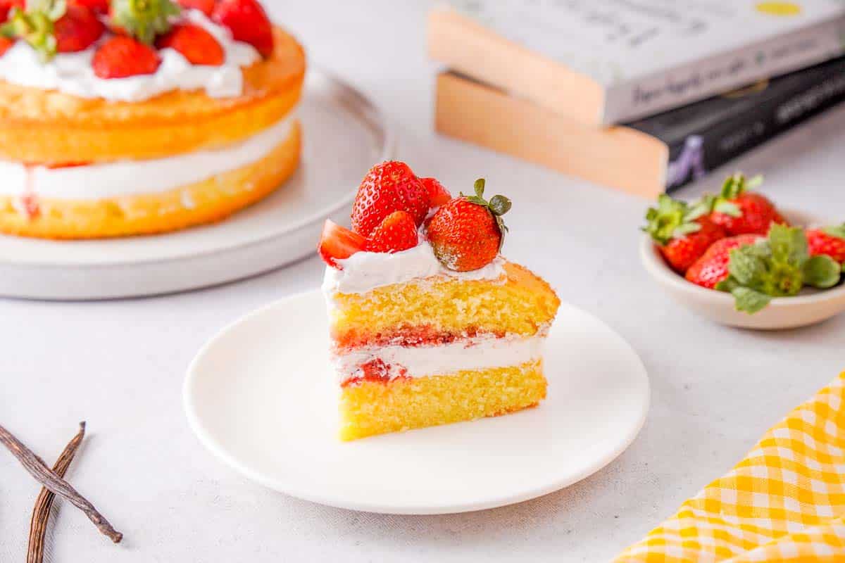 A bright photo of a slice of Victoria Sponge Cake with strawberries on top. The rest of the cake can be seen in the background.