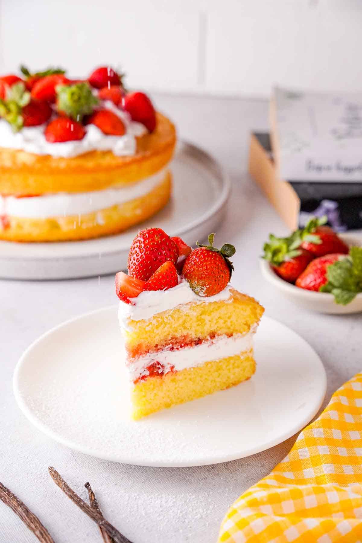 A slice of Victoria Sponge Cake with Balsamic Strawberries on a plate. The rest of the cake, as well as some additional fresh strawberries, can be seen in the background.