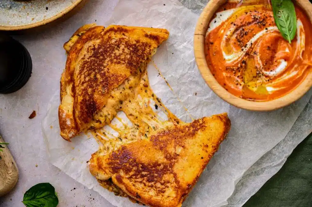 A grilled cheese sandwich with melted cheese oozing out, cut into two halves on parchment paper. Next to it is a bowl of creamy tomato soup garnished with a basil leaf and ground pepper.