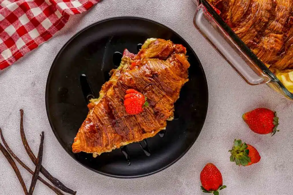 A slice of croissant pudding topped with a strawberry on a black plate. Nearby are whole strawberries, a few vanilla pods, and a red-checkered cloth. A glass dish with more pudding is partially visible.