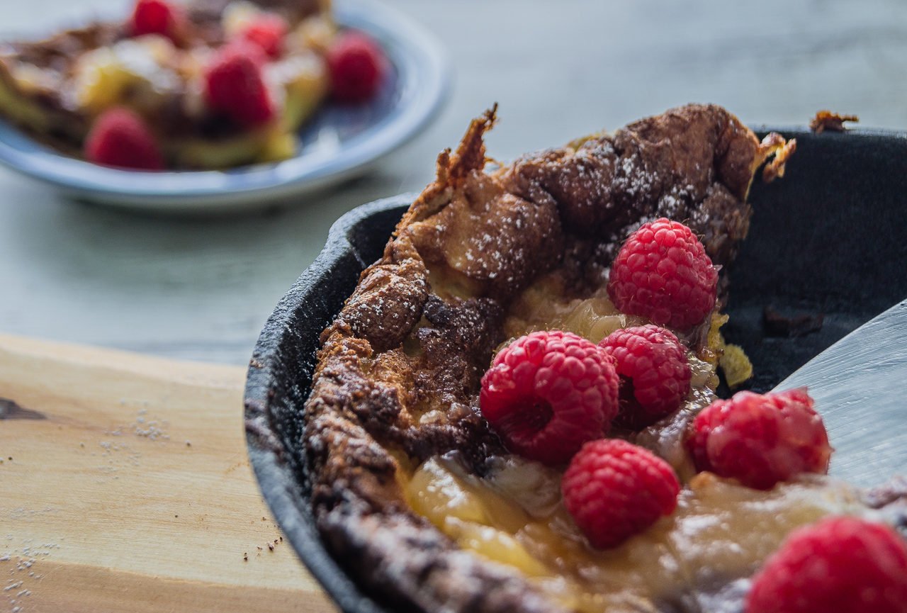 Close-up photo of Lemon Raspberry Dutch Baby Pancake with a slice cut out. The slice is seen on a plate in the background. 